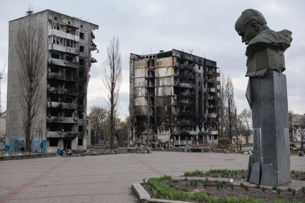 Damaged buildings with charred facades stand behind a somber statue in an empty plaza.