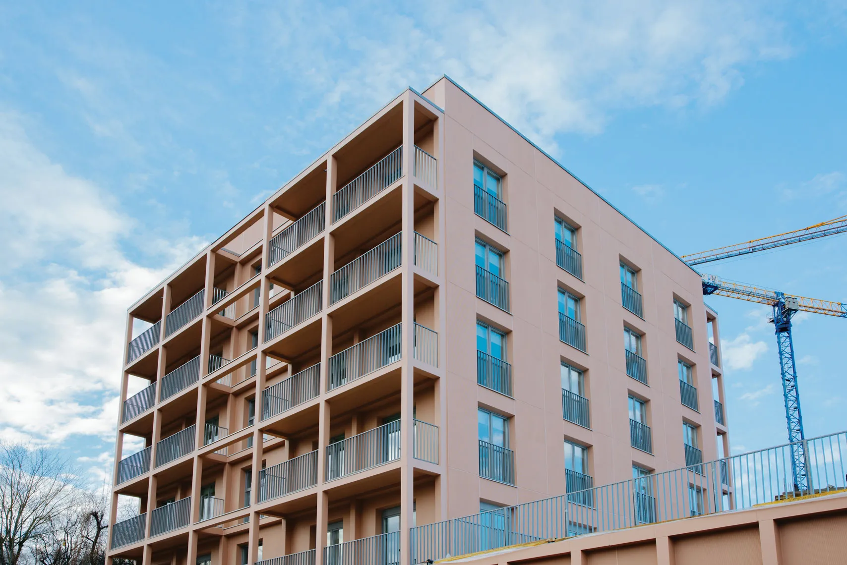 Modern peach-colored apartment building with balconies under a blue sky. A crane is visible, suggesting ongoing construction.