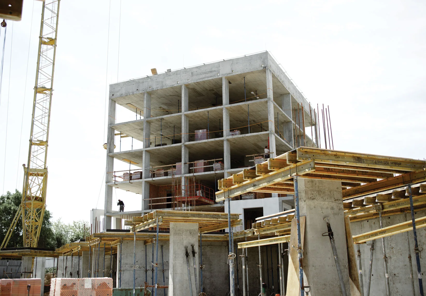 A multi-story concrete building under construction, featuring scaffolding and a crane on the left, and a construction worker visible on one of the levels.