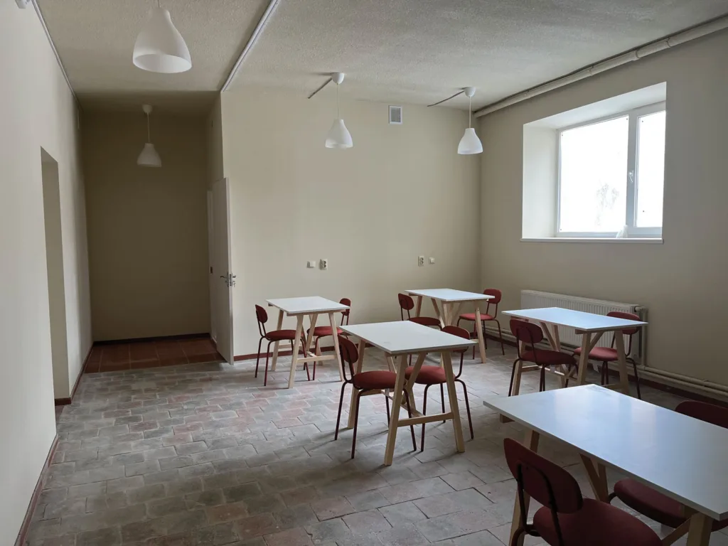 Spacious room with four white tables and red chairs, brick floor, beige walls, large window, and ceiling lights.