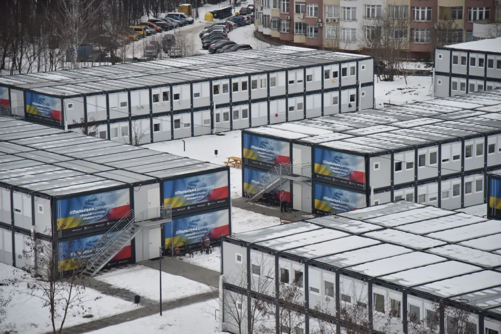 Snow-covered modular structures are arranged in rows, sides adorned with colorful banners, surrounded by snow.
