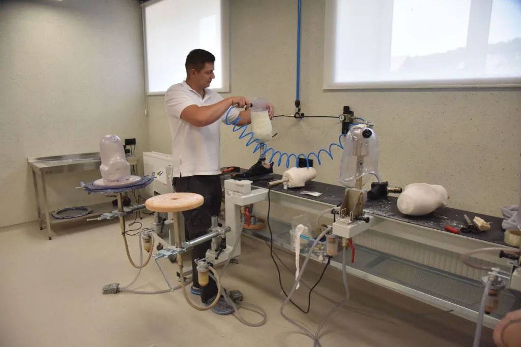 A man in a workshop is adjusting prosthetic limbs on a workbench, surrounded by tools and equipment.