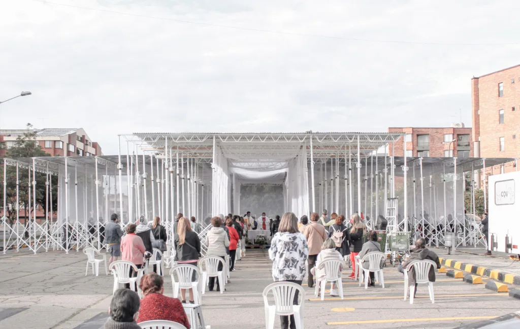 People standing and sitting around a temporary outdoor chapel made of scaffolding and drapery.