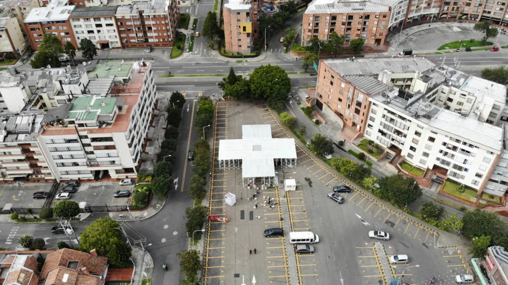 Aerial view of cross-shaped white pavilion in parking lot in the middle of a city.