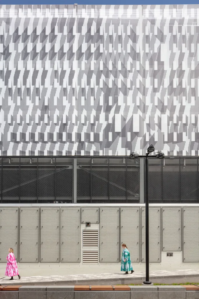 A patterned scrim wraps a concrete garage while two women in patterned dresses walk below.