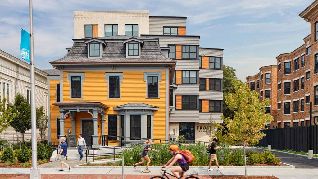 Yellow house with new addition rising behind it on a vibrant urban street.