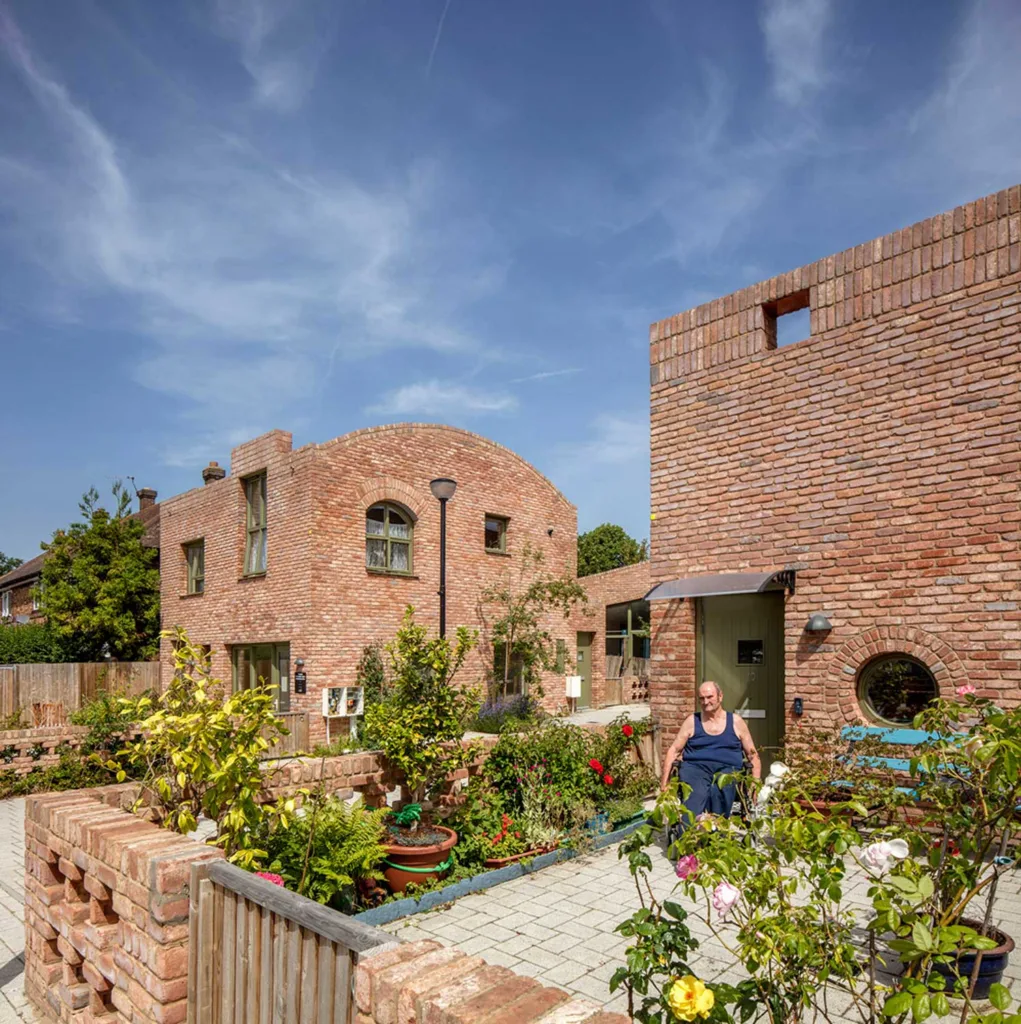 Man in wheelchair surrounded by plants on terrace of low-rise brick building.