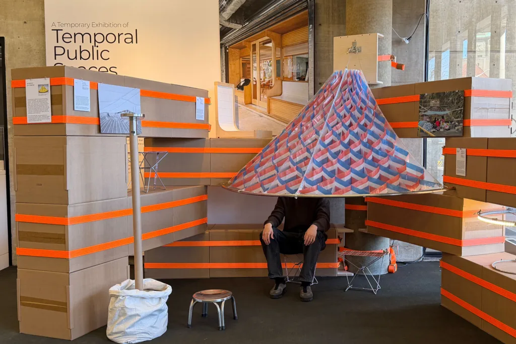 A figure sits below a red, white, and blue parasol in the exhibition "A Temporal Exhibition of Temporary Public Spaces"