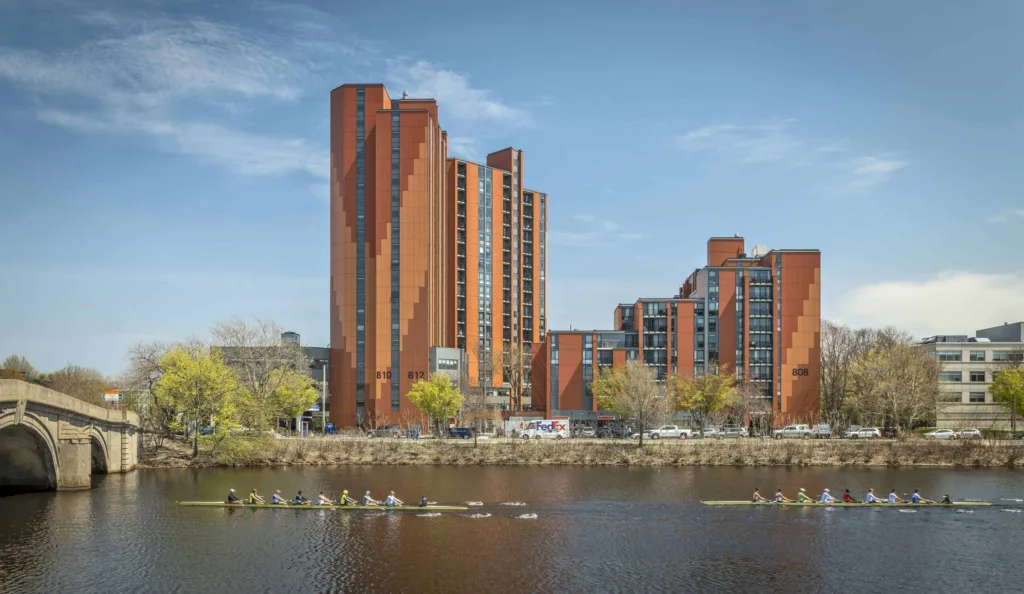 Photograph of tall terra cotta apartment building behind rowers on the Charles river.