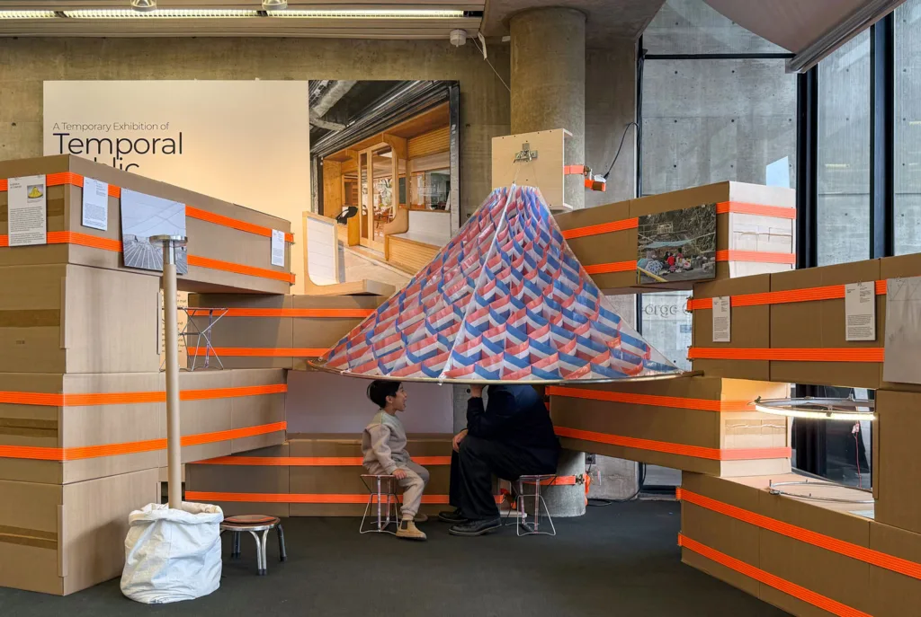 Two figures sits below a red, white, and blue parasol in the exhibition "A Temporal Exhibition of Temporary Public Spaces"