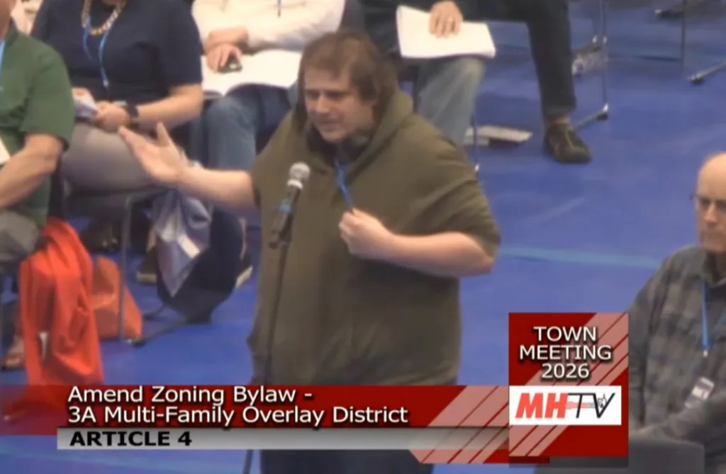 Man in dark green hoodie speaking into a microphone at a town meeting.