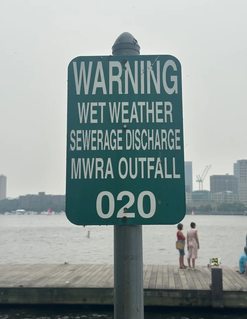 Green sign on bank of river that says "Warning Wet Weather Sewage Discharge MWRA Outfall 020"
