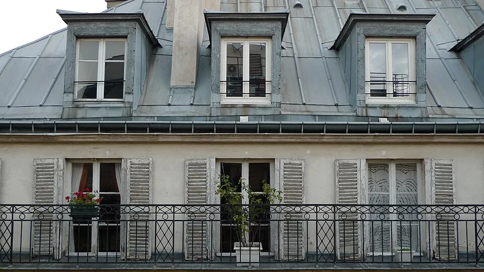 Three gabled windows jut out from the top story of a Parisian residential building, above three large windows with open shutters, a fenced balcony, and potted plants.