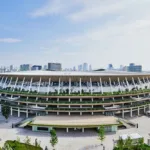 Aerial view of a large landscaped oval stadium with a city skyline in the background.