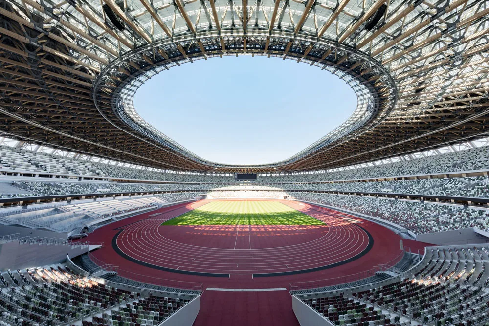 Interior of a large stadium with a red track, green field, patterned seating, and an open oval roof showing blue sky.