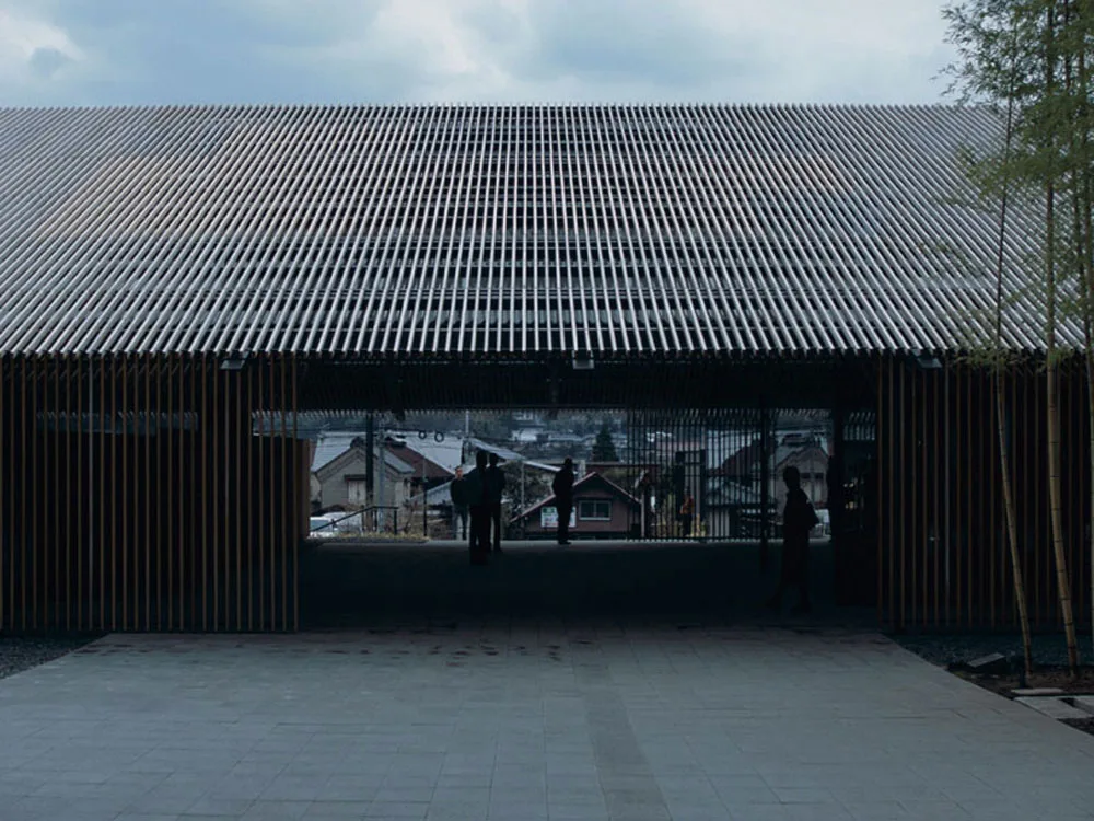 Modern slatted-roof pavilion with silhouetted people, a paved plaza, and hillside houses visible through its open center.