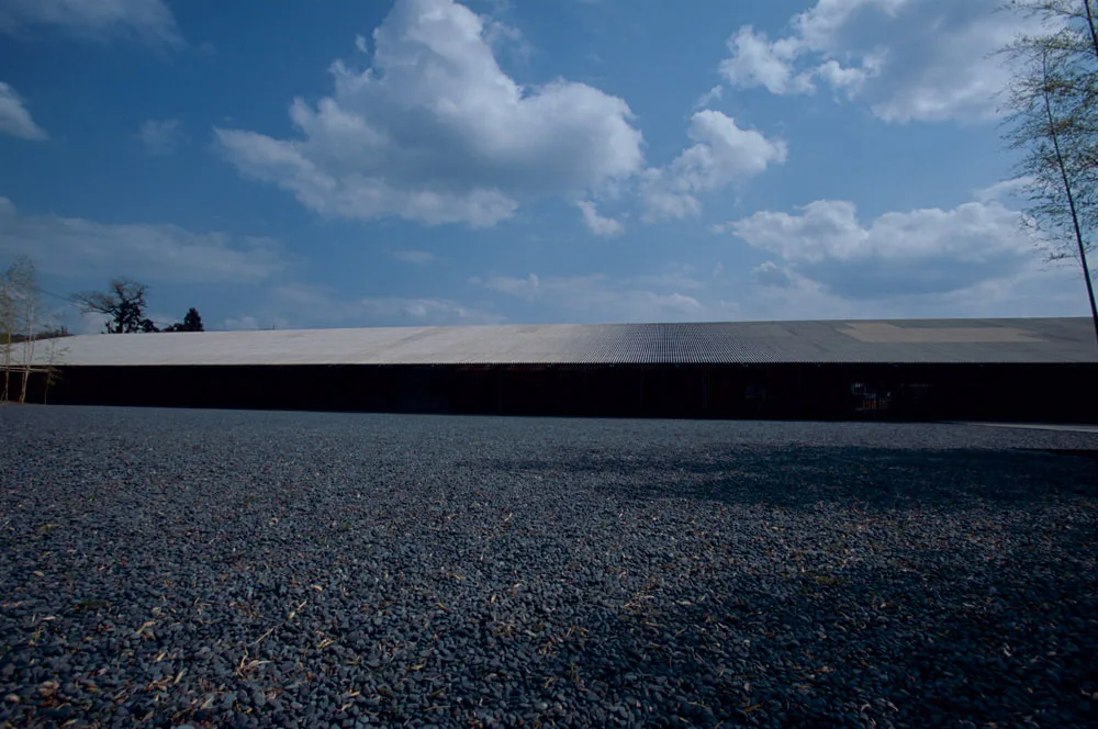 Long, low industrial building with a corrugated roof behind a broad gravel-covered foreground under a bright, cloud-filled blue sky.