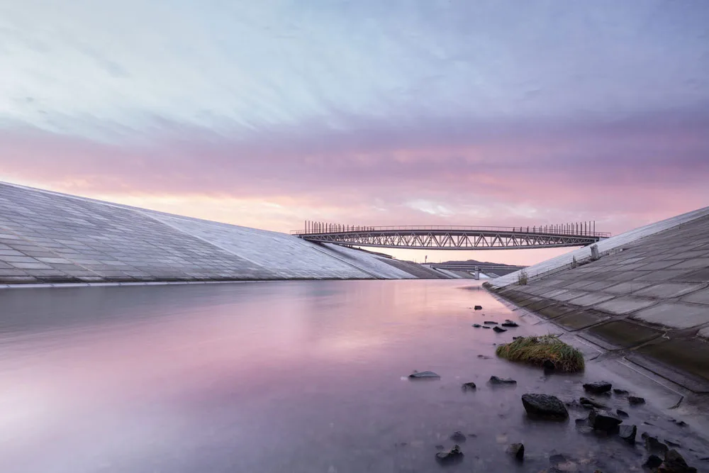 Calm canal with sloped concrete embankments and a metal truss bridge beneath a pink and lavender evening sky.
