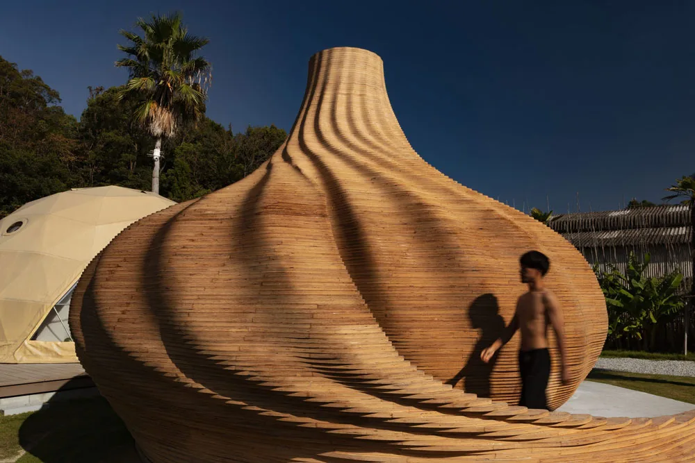 Large layered wooden sculpture with curved ridges outdoors, a palm tree behind it, and a blurred person walking in front.