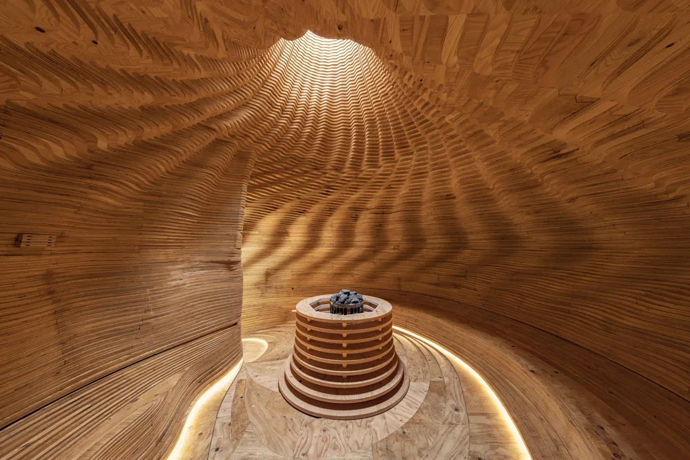 Circular wooden room with rippling walls and ceiling, a bright overhead opening, and a tiered central basin filled with dark stones.