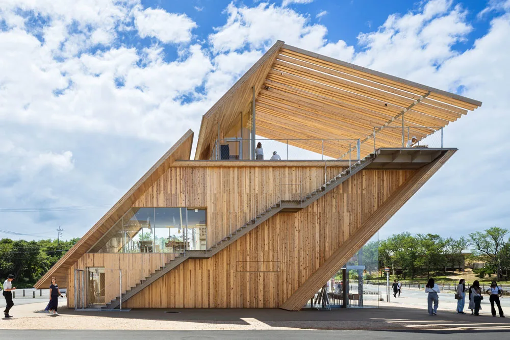 Angular wooden building with diagonal exterior staircases, glass walls, and a covered rooftop terrace under a partly cloudy blue sky.