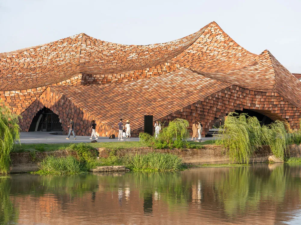 Sculptural shingle-clad building with dramatically undulating roofs beside a reflective pond.