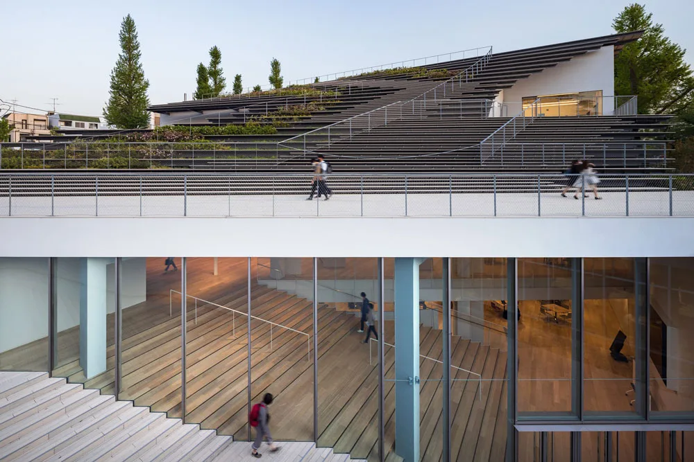 Modern multi-level building with stepped landscaped terraces, broad stairways, glass walls, and people walking inside and outside.