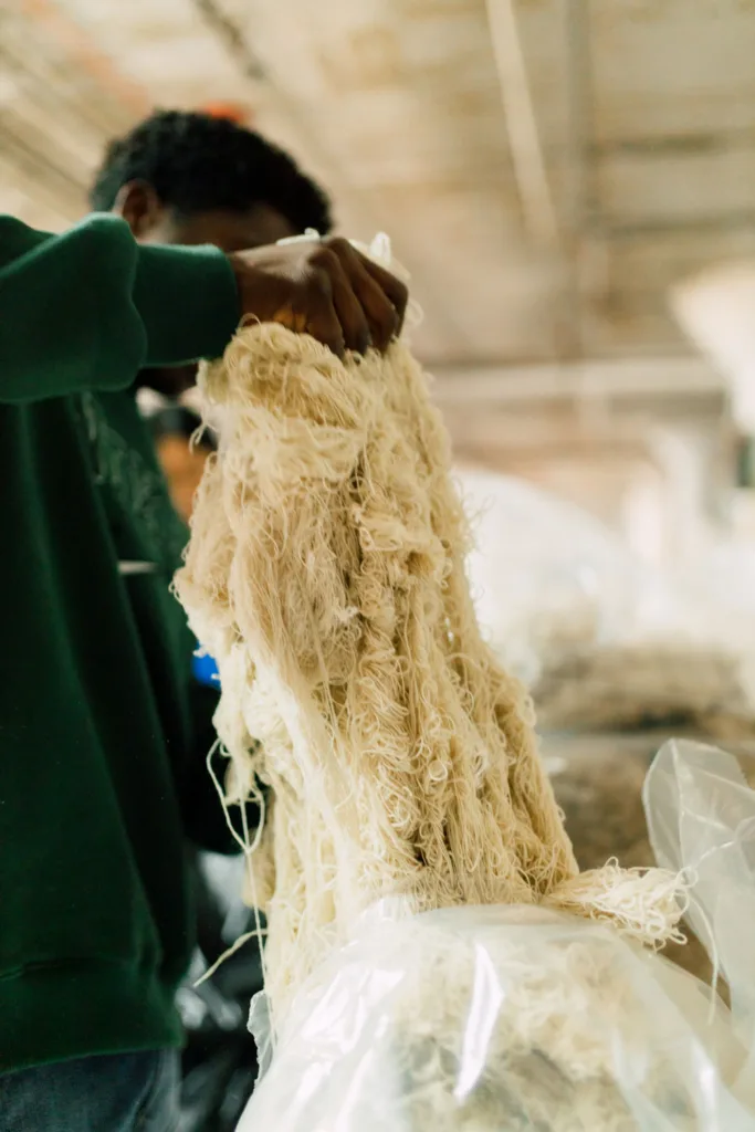 A man in a green shirt and pants pulls strands of wool from a plastic bag in a mill.