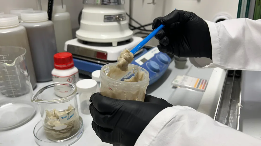 Two black-gloved Hans at a chemist's desk working with a tan, paste-like substance.