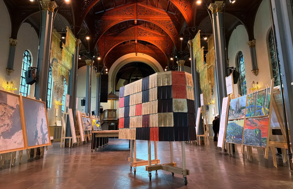 A wooden c-shaped structure sheathed in colorful shingles, sitting inside a display hall with other works.