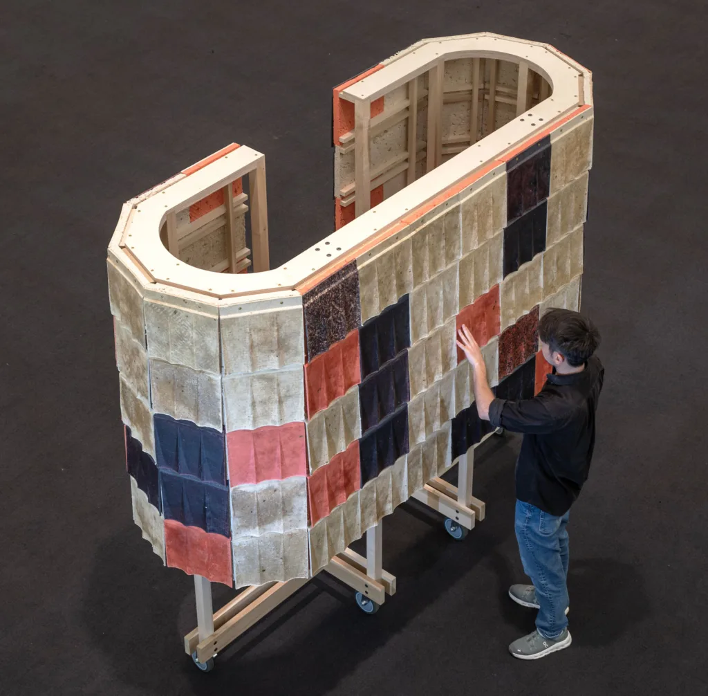 A man in a gray shirt inspects an open wooden framework that curves into a "c" shape, with insulation panels outside the framework, all covered by multicolored shingles.