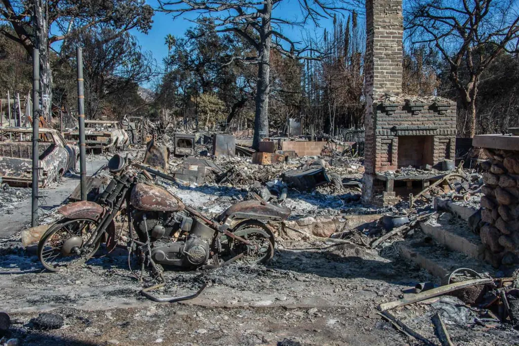 The charged remains of a neighborhood burnt by wildfires. A rusted motorcycle and lone standing hearth are the main foci of the image.