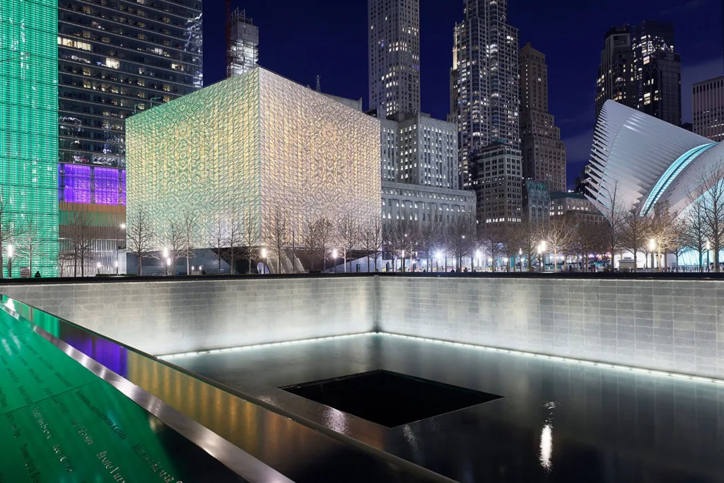 Glowing cube-shaped building behind a reflecting pond and in front of an urban skyline.