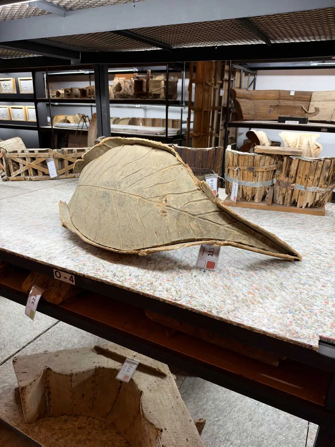 A wood mold of a large leave rests of the floor in front of other molds, stored behind on shelves.